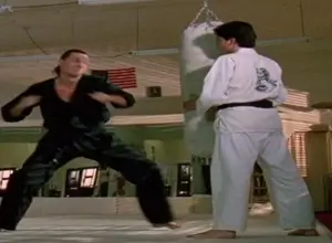 A screenshot of Terry Silver demonstrating rule 2 of the method to Daniel whilst having him hold the punching bag in the dojo whilst he strikes it